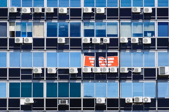 Rows of rectangular windows on a building facade, each equipped with air conditioning units. A sign with text in Spanish appears on one of the windows, indicating a space for rent. The windows vary in shades of blue, adding a uniform and repetitive architectural pattern.
