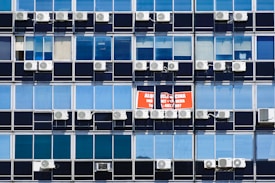 Rows of rectangular windows on a building facade, each equipped with air conditioning units. A sign with text in Spanish appears on one of the windows, indicating a space for rent. The windows vary in shades of blue, adding a uniform and repetitive architectural pattern.