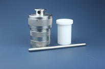 A metallic industrial component with a cylindrical shape sits alongside a white plastic container and a slender metal rod. The metallic component has a textured surface and a small hole on top. The background is a solid, muted blue.