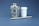 A metallic industrial component with a cylindrical shape sits alongside a white plastic container and a slender metal rod. The metallic component has a textured surface and a small hole on top. The background is a solid, muted blue.