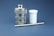 A metallic industrial component with a cylindrical shape sits alongside a white plastic container and a slender metal rod. The metallic component has a textured surface and a small hole on top. The background is a solid, muted blue.