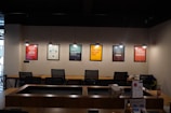 A modern office space is decorated with motivational posters on the wall. The posters are illuminated by individual hanging lights, and a long wooden desk is situated below with several office chairs. Some office supplies, a telephone, and a calendar are on the desk.