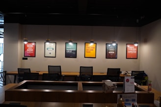 A modern office space is decorated with motivational posters on the wall. The posters are illuminated by individual hanging lights, and a long wooden desk is situated below with several office chairs. Some office supplies, a telephone, and a calendar are on the desk.