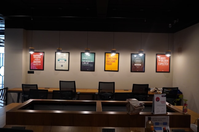 A modern office space is decorated with motivational posters on the wall. The posters are illuminated by individual hanging lights, and a long wooden desk is situated below with several office chairs. Some office supplies, a telephone, and a calendar are on the desk.