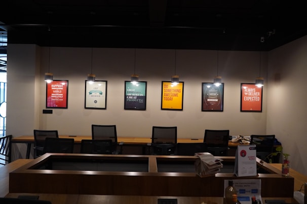 A modern office space is decorated with motivational posters on the wall. The posters are illuminated by individual hanging lights, and a long wooden desk is situated below with several office chairs. Some office supplies, a telephone, and a calendar are on the desk.