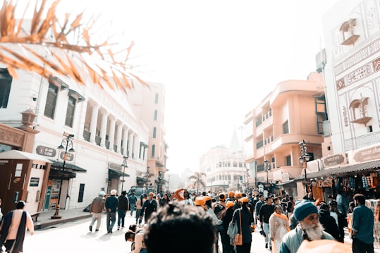 A bustling street scene filled with people, some wearing traditional clothing and turbans, walking and interacting. The architecture includes historic buildings with intricate designs and modern shops. The bright sunlight casts strong shadows, creating a sharp contrast.