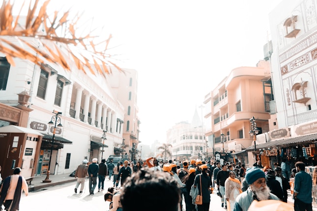 A bustling street scene filled with people, some wearing traditional clothing and turbans, walking and interacting. The architecture includes historic buildings with intricate designs and modern shops. The bright sunlight casts strong shadows, creating a sharp contrast.