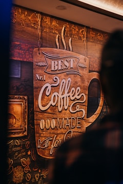 A richly decorated wooden wall features an artistic coffee-themed design. A large sign in the shape of a coffee cup displays the text 'Best No. 1 Coffee Made for You.' Surrounding the sign are intricate designs, including coffee beans and floral patterns. A framed picture hangs on the left, adding to the cozy, artisanal atmosphere.