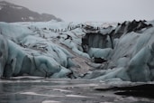 A rugged icy terrain with large crevasses and formations, featuring a line of people hiking across the glacier. The scene is set in overcast conditions, with a backdrop of snow-covered mountains.