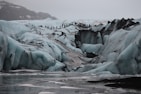 A rugged icy terrain with large crevasses and formations, featuring a line of people hiking across the glacier. The scene is set in overcast conditions, with a backdrop of snow-covered mountains.