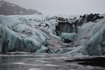 A rugged icy terrain with large crevasses and formations, featuring a line of people hiking across the glacier. The scene is set in overcast conditions, with a backdrop of snow-covered mountains.