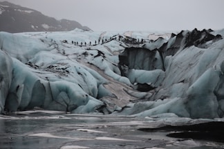 A rugged icy terrain with large crevasses and formations, featuring a line of people hiking across the glacier. The scene is set in overcast conditions, with a backdrop of snow-covered mountains.