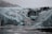 A rugged icy terrain with large crevasses and formations, featuring a line of people hiking across the glacier. The scene is set in overcast conditions, with a backdrop of snow-covered mountains.