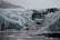 A rugged icy terrain with large crevasses and formations, featuring a line of people hiking across the glacier. The scene is set in overcast conditions, with a backdrop of snow-covered mountains.