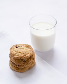 A stack of three chocolate chip cookies is placed on a white napkin next to a clear glass filled with milk. The scene is minimalist and bright, with the cookies displayed prominently against a simple backdrop.