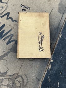A worn, vintage book with a sketch of a boy walking away on the cover lies on a textured, grey surface with graffiti-like designs, including the words 'BOOBS ON'.
