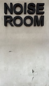 Large black 3D letters spell out 'NOISE ROOM' against a plain, slightly dirty beige wall.