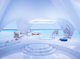 A modern, minimalist room opens up to a stunning ocean view. The space features sleek, curved architecture with white textured ceilings and marble floors. There is a small circular pool surrounded by cozy seating including a lounger, a hanging chair, and a cushioned seat. A sofa with green accent pillows, a table with snacks, and a jug of refreshing drink complete the serene, luxurious setting.