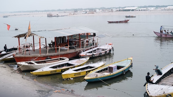 Several small wooden boats are docked along a sandy riverbank. A larger boat with a canopy is anchored nearby, serving as a floating platform or shop. People are sitting on the boats and one person is fishing further out on the river. The background features a wide expanse of water, with more boats moving in the distance, and a hazy shoreline is visible.