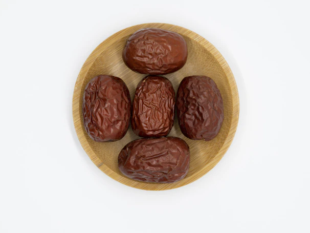 Five dried, wrinkled brown dates are arranged on a round, light-colored wooden plate set against a plain white background.