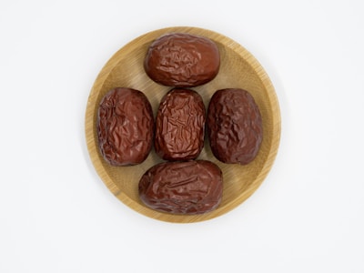 Five dried, wrinkled brown dates are arranged on a round, light-colored wooden plate set against a plain white background.