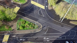 Aerial view of a multi-lane roundabout intersection featuring several crosswalks with bold yellow and white stripes. Surrounding the intersection are patches of green grass and trees, along with some paved pathways. Traffic signs are visible along the roads, and the entire area is well-marked with road lines and arrows.
