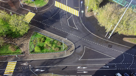 Aerial view of a multi-lane roundabout intersection featuring several crosswalks with bold yellow and white stripes. Surrounding the intersection are patches of green grass and trees, along with some paved pathways. Traffic signs are visible along the roads, and the entire area is well-marked with road lines and arrows.