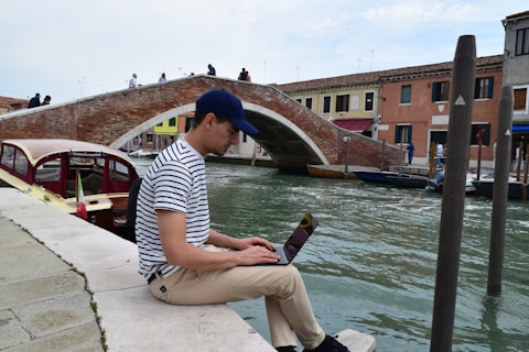 A person wearing a striped shirt and a cap sits on a stone edge by a canal, using a laptop. In the background, a brick arched bridge spans across the canal with a few people crossing it. On the canal, a boat with an Italian flag is visible, and buildings line the opposite side of the water.