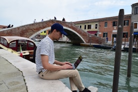 A person wearing a striped shirt and a cap sits on a stone edge by a canal, using a laptop. In the background, a brick arched bridge spans across the canal with a few people crossing it. On the canal, a boat with an Italian flag is visible, and buildings line the opposite side of the water.