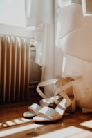 Strappy high-heeled sandals are placed on a wooden floor next to a white dress. Soft natural light streams in from a nearby window, casting gentle shadows.