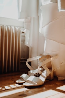 Strappy high-heeled sandals are placed on a wooden floor next to a white dress. Soft natural light streams in from a nearby window, casting gentle shadows.