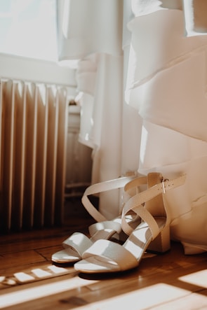 Strappy high-heeled sandals are placed on a wooden floor next to a white dress. Soft natural light streams in from a nearby window, casting gentle shadows.