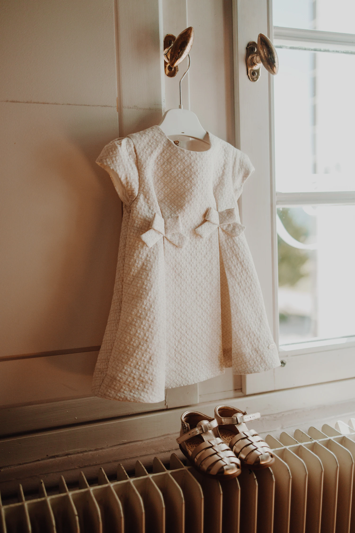 Cream baby dress hanging in soft window light with tiny shoes below
