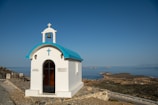A small white chapel with a blue roof sits on a rocky hill overlooking a vast expanse of ocean. The chapel features a cross and bell on top, and there are signs on either side of the entrance. The surrounding landscape includes rugged terrain and distant islands on a clear day.