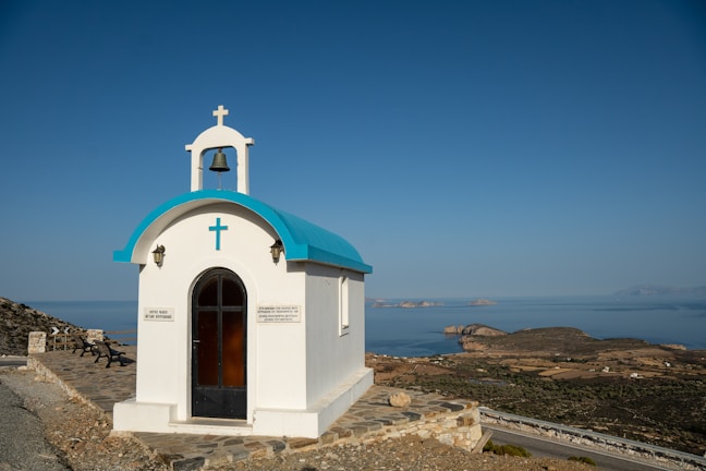A small white chapel with a blue roof sits on a rocky hill overlooking a vast expanse of ocean. The chapel features a cross and bell on top, and there are signs on either side of the entrance. The surrounding landscape includes rugged terrain and distant islands on a clear day.