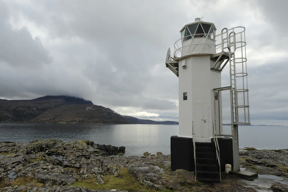 The image depicts a serene coastal scene featuring a lighthouse standing tall on a rocky outcropping, overlooking the vast expanse of water below. The lighthouse is painted white and has a black railing around its base for safety purposes. A small staircase leads up to the entrance of the lighthouse, inviting visitors to explore the nautical world beyond.

The sky above is filled with clouds, casting a soft light over the scene. In the distance, mountains can be seen, adding depth and grandeur to the landscape. The water below mirrors the cloudy sky, creating a beautiful reflection that enhances the overall ambiance of the image.