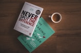 Two books are placed on a wooden surface beside a cup of coffee. One book has a white cover with the title 'Never Split the Difference' in bold red and black text. The other book has a turquoise cover with the title 'The Making of a Manager' in white text.