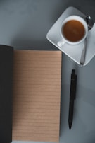 An open notebook and pen beside a cup of coffee on a wooden table.