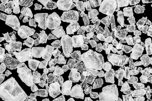 A collection of irregular, translucent crystals is scattered over a dark background. The crystals vary in size and shape, with a few appearing larger and more prominent than others. Their texture looks rough and some have a shimmering effect, indicating light reflection.