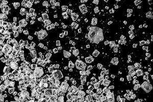 Numerous translucent crystals, likely salt or sugar, are scattered on a black background. The crystals vary in size and shape with some appearing cubic and others more irregular. The contrast between the dark background and the sparkling crystals highlights their clear, multifaceted texture.
