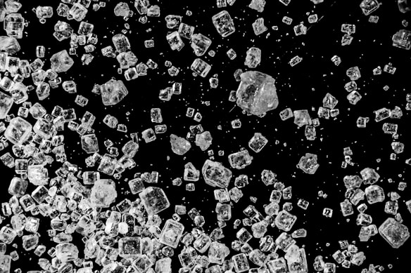 Numerous translucent crystals, likely salt or sugar, are scattered on a black background. The crystals vary in size and shape with some appearing cubic and others more irregular. The contrast between the dark background and the sparkling crystals highlights their clear, multifaceted texture.