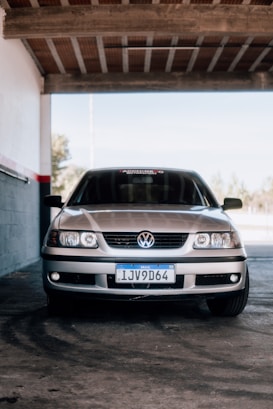 A silver Volkswagen car is parked inside a shaded garage area. The front view highlights the vehicle's headlights, grille, and license plate.
