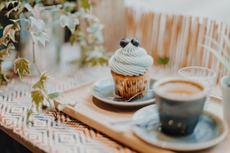 A cupcake with light blue frosting topped with blueberries is placed on a small plate with a spoon beside it. It sits on a wooden tray alongside a cup of coffee and a glass of water. Leafy plants surround the arrangement, adding a natural touch to the setting.