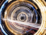 A dynamic and abstract display of circular light trails creating a vortex effect. The image features a swirling pattern with golden, white, and blue lights against a dark background, suggesting motion and energy.
