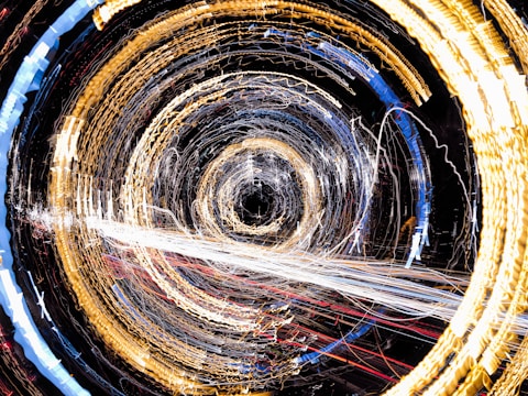 A dynamic and abstract display of circular light trails creating a vortex effect. The image features a swirling pattern with golden, white, and blue lights against a dark background, suggesting motion and energy.