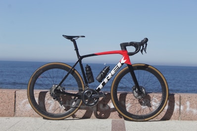 A sleek road bicycle with a predominantly black frame accented by a striking red and white design is positioned against a stone barrier. The bicycle features aerodynamic racing handlebars, a water bottle, and high-performance tires. The background showcases a serene ocean view under a clear blue sky, giving the scene an open and expansive feel.
