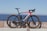 A sleek road bicycle with a predominantly black frame accented by a striking red and white design is positioned against a stone barrier. The bicycle features aerodynamic racing handlebars, a water bottle, and high-performance tires. The background showcases a serene ocean view under a clear blue sky, giving the scene an open and expansive feel.