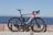 A sleek road bicycle with a predominantly black frame accented by a striking red and white design is positioned against a stone barrier. The bicycle features aerodynamic racing handlebars, a water bottle, and high-performance tires. The background showcases a serene ocean view under a clear blue sky, giving the scene an open and expansive feel.