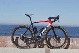 A sleek road bicycle with a predominantly black frame accented by a striking red and white design is positioned against a stone barrier. The bicycle features aerodynamic racing handlebars, a water bottle, and high-performance tires. The background showcases a serene ocean view under a clear blue sky, giving the scene an open and expansive feel.