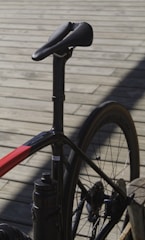 A close-up view of a bicycle focusing on the seat, seat post, and rear section of the frame and wheel. The bicycle has a sleek, modern design with a predominantly black frame and red accents. The background consists of wooden planks creating a rustic setting.